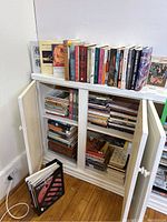 Books and magazines arranged in a white cabinet with doors open, some books stacked inside and a few on top of the unit.