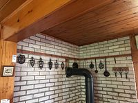 Wide shot of wall display showing seven cast iron trivets and three ladles hung above a stove pipe.