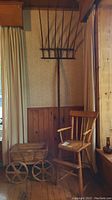 Full view of the three items grouped together: spindle back doll chair, wooden wagon with large wheels, and tall wooden pitchfork against a wood-paneled wall and curtain.
