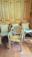 Four rattan chairs placed in front of wooden cupboard, three with cushions and one without.