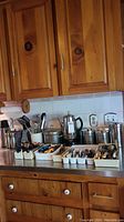Wide view of kitchen counter showing trays of assorted cutlery, metal canisters, coffee percolator and other kitchenware items.