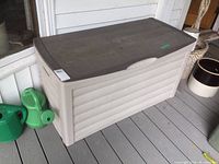 Side view of the beige plastic deck box with brown lid, showing integrated handle and adjacent green watering cans.