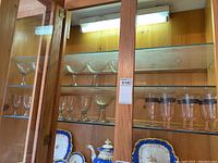 Photo showing the glassware collection with multiple clear coupe glasses with blue stems on upper shelf and pink wine glasses with black bands on rim on right side of lower shelf.