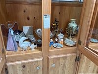 Wide shot of pine wood cabinet containing stained glass angel figurine, carved stone animals, trinket boxes, watches, vases, candleholders, and jewelry tree with earrings