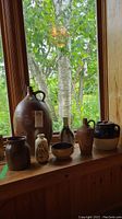 Wide view of all stoneware items on wood windowsill including large jug, bean pot with lid, smaller painted vases, dish, and pots.