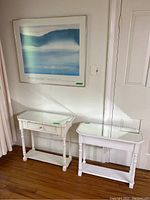 Photo of two white wooden tables with mirrored tops positioned side by side against a wall under the Pedie Wolfond poster. One table has a drawer; the other is open underneath.
