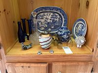 Shelf with blue and white Ridgeway Willow platter, blue and white decorative plate, ceramic ginger jar, blue glass candlesticks, blue glass bottle, aperitif glasses, decorative vase, decanter, ceramic sheep and bird figurines.