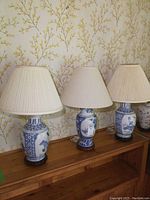 Front view of three blue and white porcelain table lamps with white fabric shades, standing on wooden surface against floral wallpaper background.
