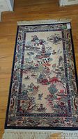 Full view of small village scene rug with fringe, laid on wood floor.