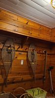 Pair of vintage wooden snowshoes hanging on a wood-paneled wall, along with two wooden canes and two metal grabbing tools leaning nearby.