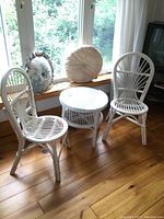 Full set of two white wicker chairs and one round wicker table with glass top on wooden floor by window, natural light.