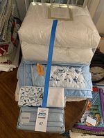 Stack of bedding items showing new mattress pad, blue floral comforter, and packaged sheet sets