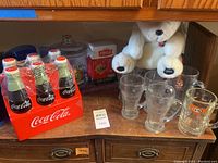 Wide view showing assembled Coca-Cola glass mugs, A&W mugs, Coca-Cola soft drink holder with sealed bottles, white plush Coca-Cola bear, Hershey's treat jar, and cookie jars arranged on counter.