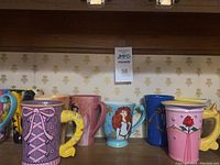 Shelf with mostly tall Disney princess mugs showing various designs including pink mugs with bow and rose, blue mugs with Merida and Beauty and the Beast text