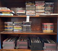 Wide shot of wooden shelving holding stacks of DVDs, Blu-rays, VHS tapes and CD storage cases. Shows the large quantity and storage method for the lot.