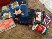 Pile of six folded handmade quilts in assorted colors and patterns, plus sewing scissors and fabric carrier bag
