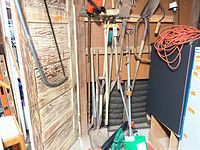 Wide shot showing wall with hanging garden tools including shovels, saw, and pruning tools next to pegboard and door
