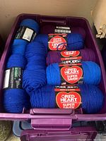 Purple bin filled with various skeins of red heart super saver acrylic yarn in blue and purple colors.