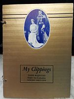 Front cover of the scrapbook in metallic gold color with an oval photo of King George VI and Queen Mother, black text title below the photo.
