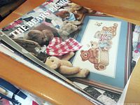 Close-up of a stack of needlepoint pattern books and leaflets focusing on teddy bear themed projects, showing titles and some project images with bears and picnic designs.