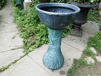 Full view of the blow mold planter on its pedestal base standing on a stone path with greenery surrounding it.