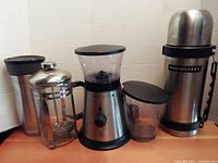 Photograph of the four coffee accessories: Contigo insulated cup, French press, electric PC grinder, and Arcosteel thermos arranged side by side on a surface.