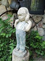 Full front view showing the girl statue standing barefoot on a stump with foliage and stone wall behind