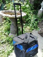 Black and blue insulated picnic bag on wheels with extended telescoping handle and carry handles in outdoor garden setting.