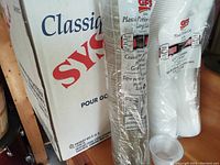 Photo showing labeled box with 'Classic Sysco' branding and a large stacked bundle of plastic portion cups with lids.