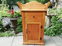 Front view of pine washstand showing its single drawer, paneled cabinet door with metal hinge and latch, and raised wooden side panels.
