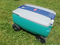 Side view of Rubbermaid cooler showing teal body, gray lid, and large black wheels on grass.
