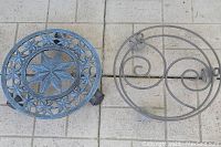 Photo of two metal plant stands side by side on tiled flooring: one ornate with star/floral pattern on wheels, other simpler circular wire stand.