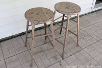 Pair of vintage metal stools with heavily worn light brown paint finish and industrial style, showing scuffs, scratches, and patina.