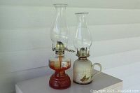Front view of two vintage oil lamps on gray table against white siding background. Left lamp is glass hurricane style with amber base, right lamp is ceramic with floral design and handle.