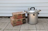 Pressure canner with two boxes of mason jars stacked beside it outdoors