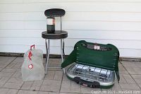 Photo showing all items in the lot: Coleman stove, propane canister on black folding stool, and two water jugs on tiled floor against white wall.