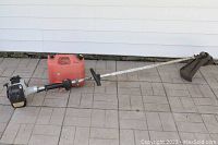 Echo SRM-261T gas/oil string trimmer with attached red jerry can placed on tiled floor, showing the full length of the tool from engine end to trimming head.