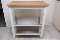 Front view of white wood kitchen cart with two metal shelves and natural wood butcher block top.