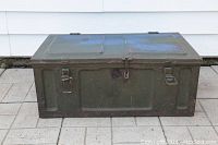 Front and top closed view of green military metal foot locker showing rectangular shape, metal latches, and blue spray paint on lid.