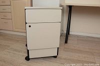Front view of the pale gray filing cabinet showing its solid rectangular shape with top drawer and larger locked drawer below, mounted on wheels.