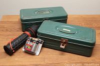 Two green metal toolboxes with handles and original labels, one stacked on the other, alongside a black and orange Garrity flashlight and a pack of Energizer D batteries on wooden surface.