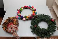 Three seasonal wreaths on white table, showing grapevine wreath with colorful pom poms, fall harvest wreath with autumn leaves, and green pine garland wreath.
