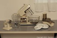 Full view of vintage Oster Kitchen Center stand mixer with glass mixing bowl, food grinder, slicer/shredder attachments, blender jar, and two beaters on the countertop.