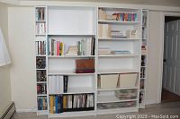 Front view of the four white melamine bookcases in a room, showing shelving filled with books and other items, hole cut-out visible on one large unit near bottom.