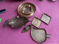 Photo of six assorted brass ashtrays including round, leaf-shaped, square, rectangular, and scoop-like pieces placed on a purple surface showing tarnish and wear.