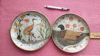 Two brass decorative plates with enameled and textured bird images, one crane and one duck, each about 8 inches in diameter with floral backgrounds.