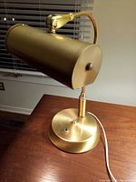 Brass table lamp with cylindrical shade on a round base, photographed indoors in front of a window blind.