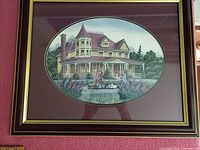 Framed 20x16 inch oval print showing a Victorian house with woman pushing a doll carriage and two dogs, under glass, with maroon matting and black/gold frame.