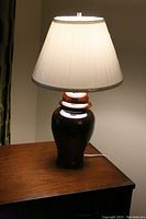 Lit view of ceramic lamp showing the beige pleated shade and brown ceramic base on a wooden surface