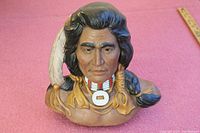 Front view of the ceramic bust showing detailed facial features, feather in hair, and necklace with pendant.